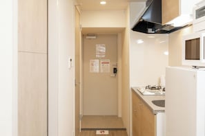A narrow hallway leading to a door with multiple signs, a kitchen area with a stove and microwave, and a wooden floor. - NOVI Dotonbori  (CHUO-KU OSAKA)