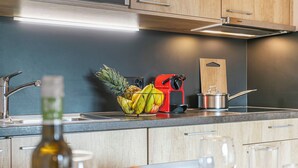 Une cuisine moderne avec un évier en acier inoxydable, un panier de fruits, un appareil électroménager rouge et une planche à découper en bois.