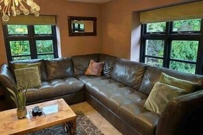 A living room with a dark leather sectional sofa, a wooden coffee table, and large windows with a view of greenery.