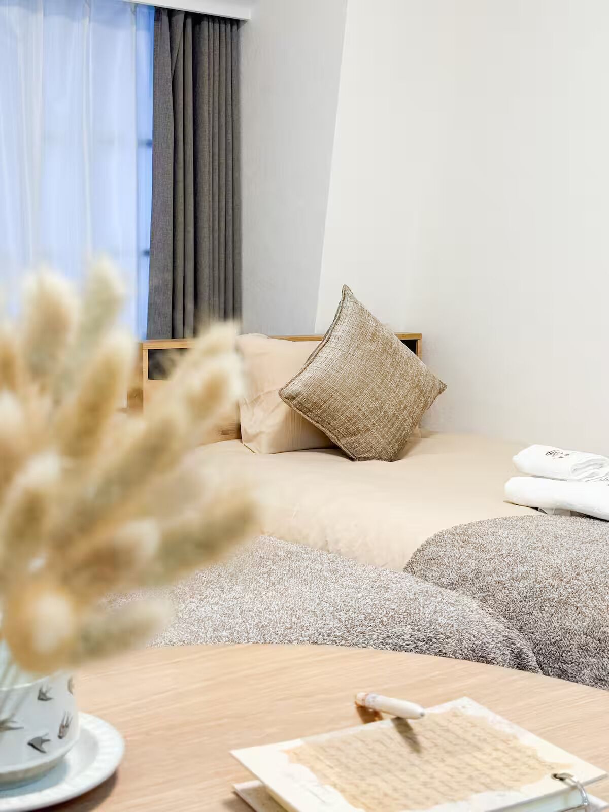 Ein Schlafzimmer mit einem Bett, Kissen, einem Nachttisch mit Buch und Stift sowie einem Fenster mit Vorhängen.