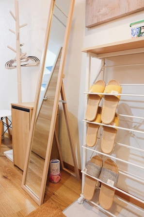 Una habitación con piso de madera, un espejo, un zapatero con pantuflas y un pequeño armario de madera.