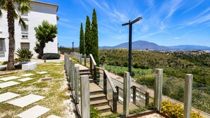 A white building with a wooden staircase leading to a balcony overlooking a landscaped area with cypress trees and a distant mountain range. - Elevated Hillside Apartment Close To Duquesa Marina (Manilva)