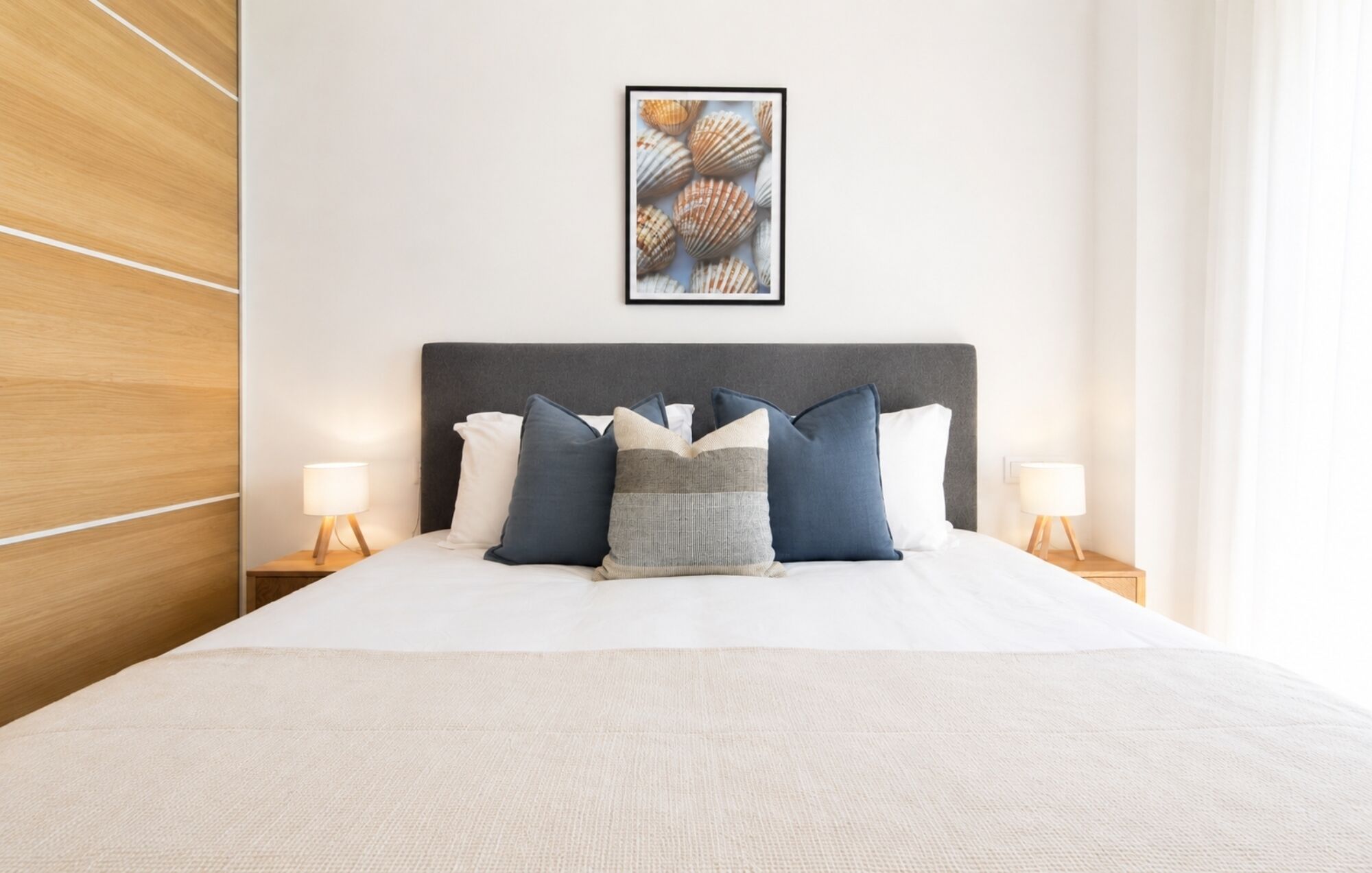 Un lit bien fait, avec une tête de lit grise, deux tables de chevet avec des lampes et, au mur, un tableau encadré représentant des coquillages.