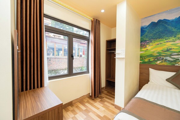 Habitación de hotel con una cama, una ventana con cortinas, un banco de madera, un cuadro en la pared y vista a un edificio.