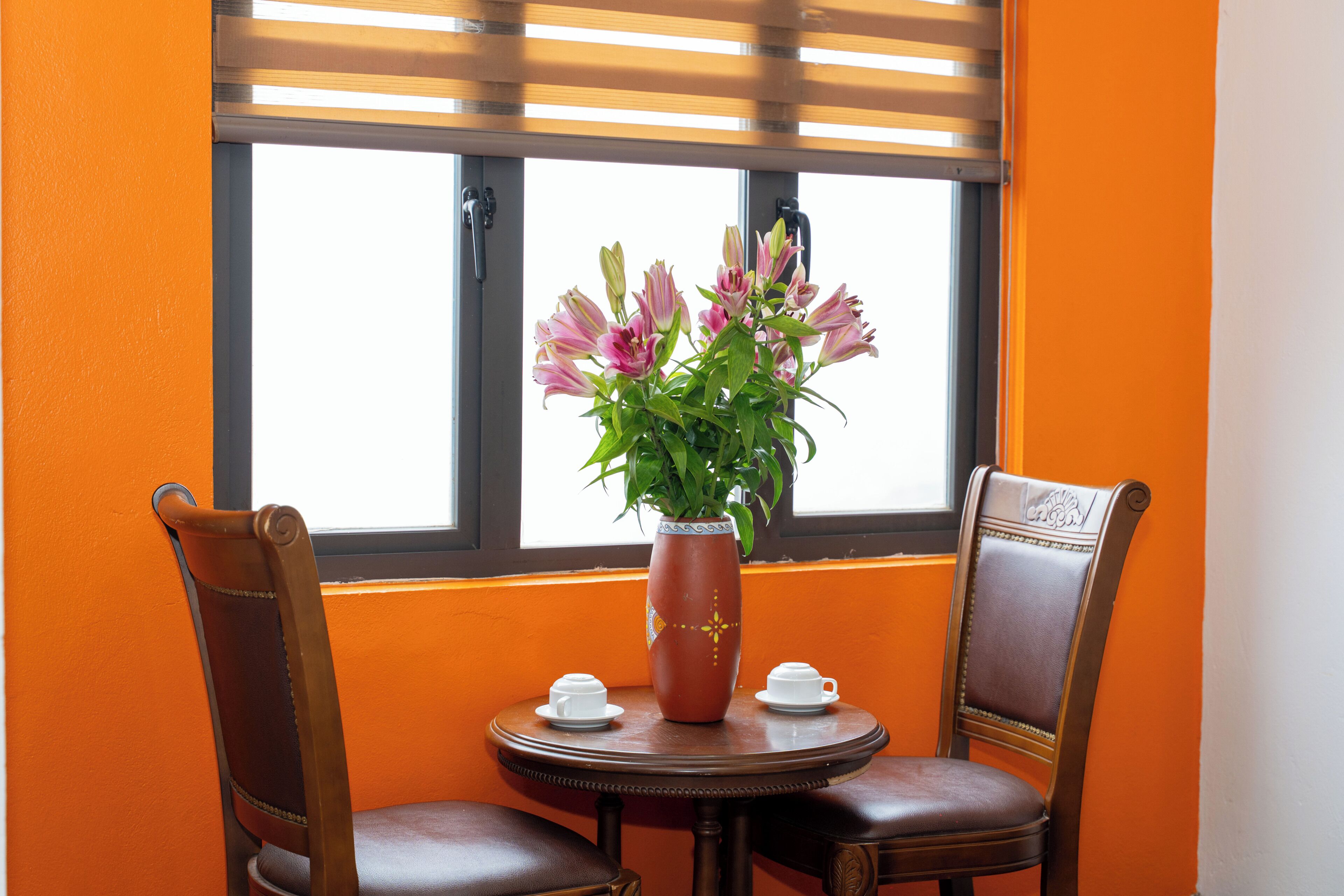 A table with a vase of flowers, two cups, and two chairs in a room with orange walls and a window.