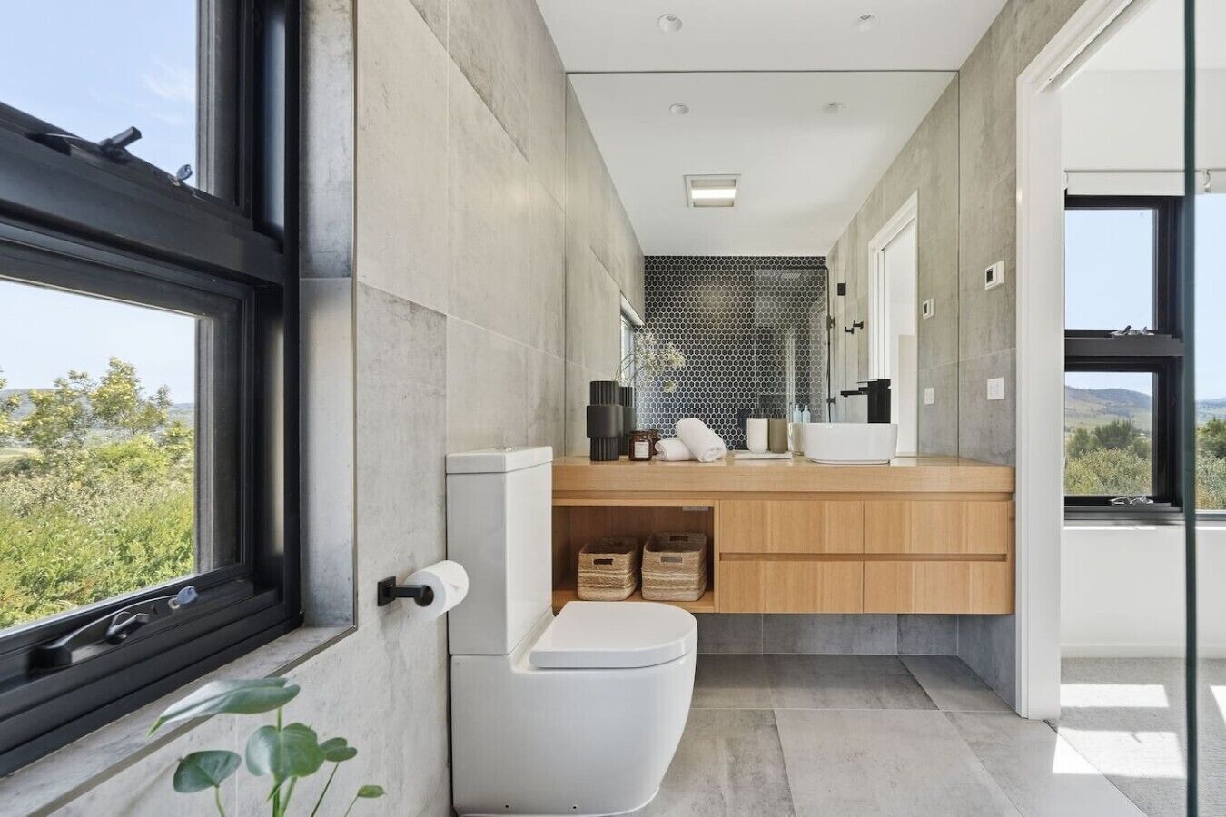 A modern bathroom with a toilet, sink, and shower area, featuring large windows and a view of the outdoors.