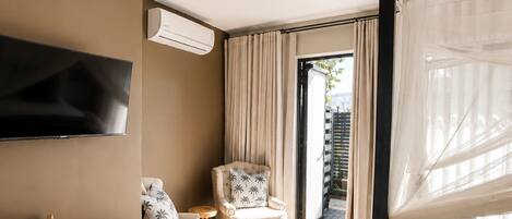 Una habitación de hotel moderna con una televisión de pantalla plana, una mesa de comedor de madera, un sillón cómodo y un balcón con cortinas.