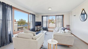 A spacious living room with two sofas, a small round table, and a television. Large windows with curtains provide natural light and views.