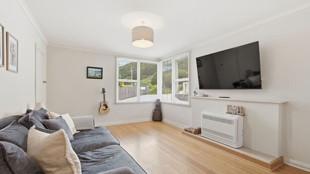 Ein modernes Wohnzimmer mit einem grauen Sofa, einer Gitarre, einem Fernseher und einem Fenster mit Jalousien.