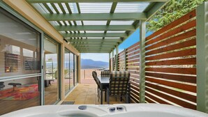 A hot tub with a view of mountains, a patio with a table and chairs, and a wooden fence. - Riverview Townhouse Retreat (Lindisfarne)
