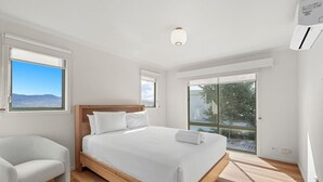 A modern bedroom with a wooden bed, white bedding, a white armchair, and large windows offering a view of the outdoors. - Riverview Townhouse Retreat (Lindisfarne)