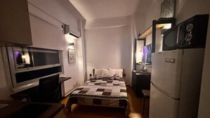 A small, compact room with a bed, a desk, a chair, a television, and a refrigerator. - Modern One-Bedroom Apartment in Kypseli - 1000 Mbps Fiber Wi-Fi (Athina)