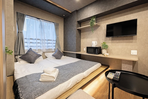 Ein modernes Hotelzimmer mit Bett, Nachttisch, Fernseher und einer kleinen Ablagefläche mit einer Pflanze.