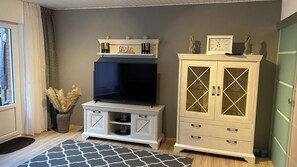 A living room with a flat-screen TV on a stand, a cabinet with glass doors, and a patterned rug. - Cozy Vacation Rental in Butjadingen in a Quiet Setting (Butjadingen)