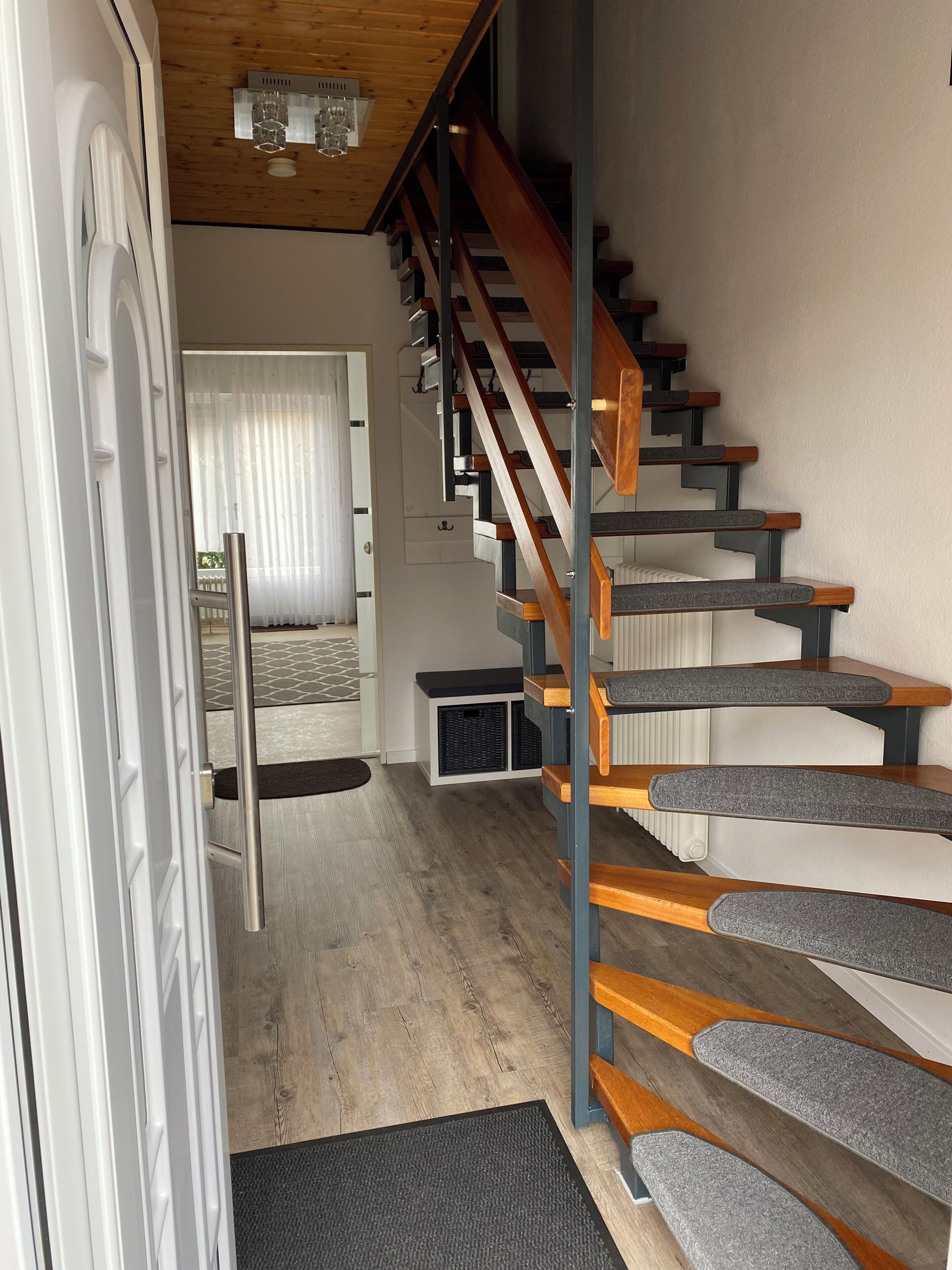 A modern interior with a wooden staircase, a white door, and a view into a room with a bed and curtains.