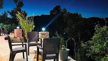 An outdoor seating area with wicker chairs and a stone fireplace, surrounded by greenery.