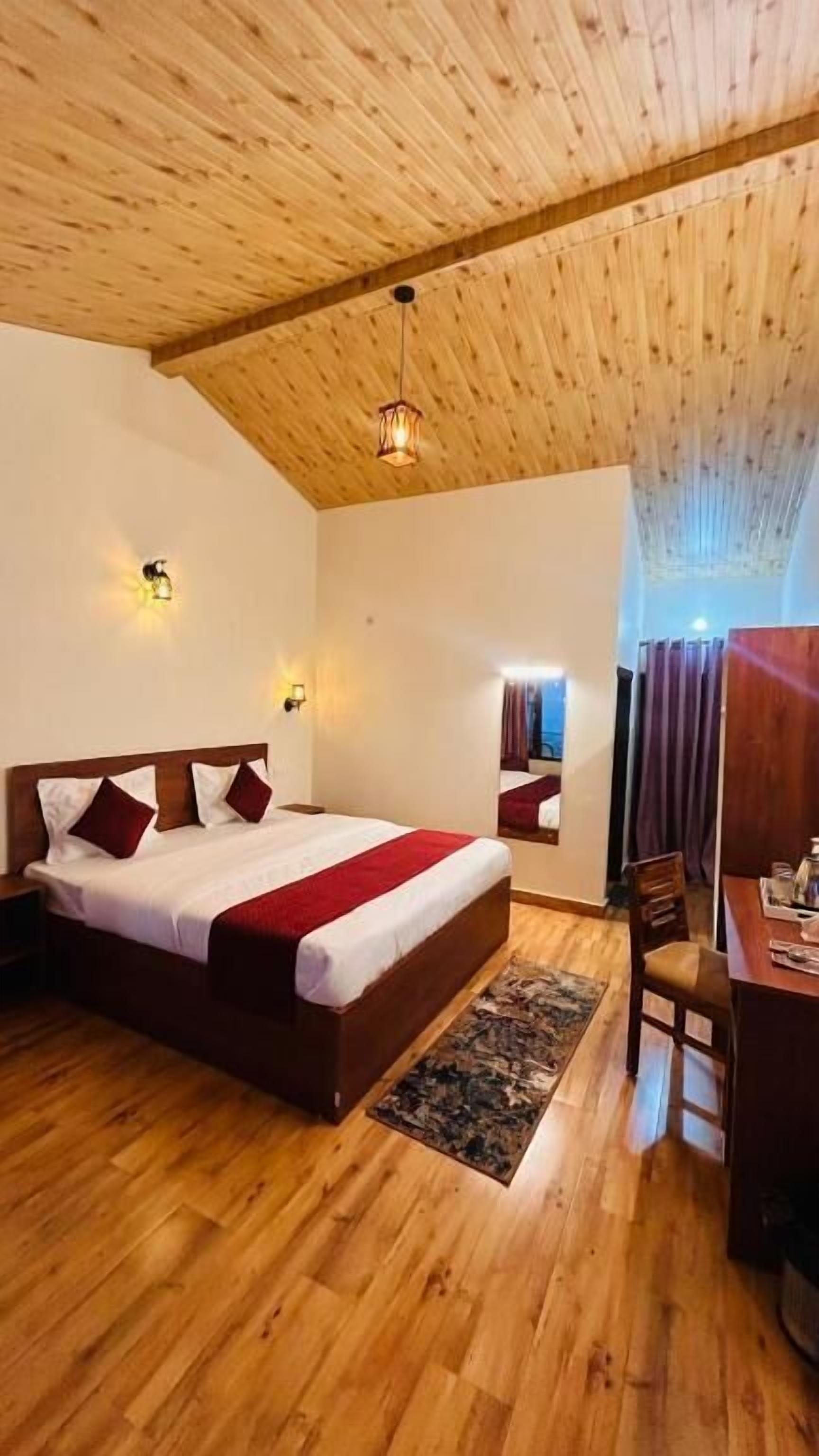 Een houten plafond, een bed met een rode deken, een bureau met een stoel en een vloerkleed.