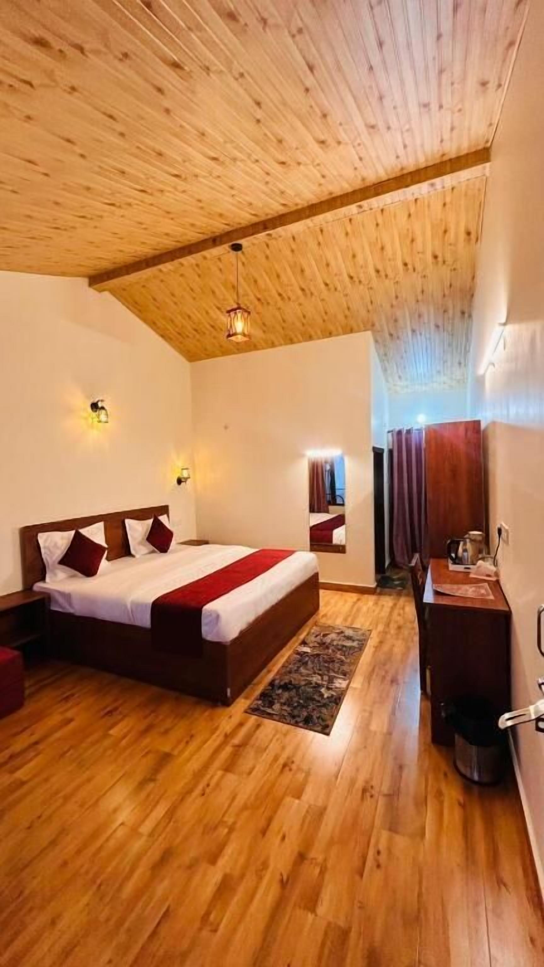 Un dormitorio con techo de madera, una cama con colcha roja y blanca, suelo de madera y un escritorio pequeño con una silla.