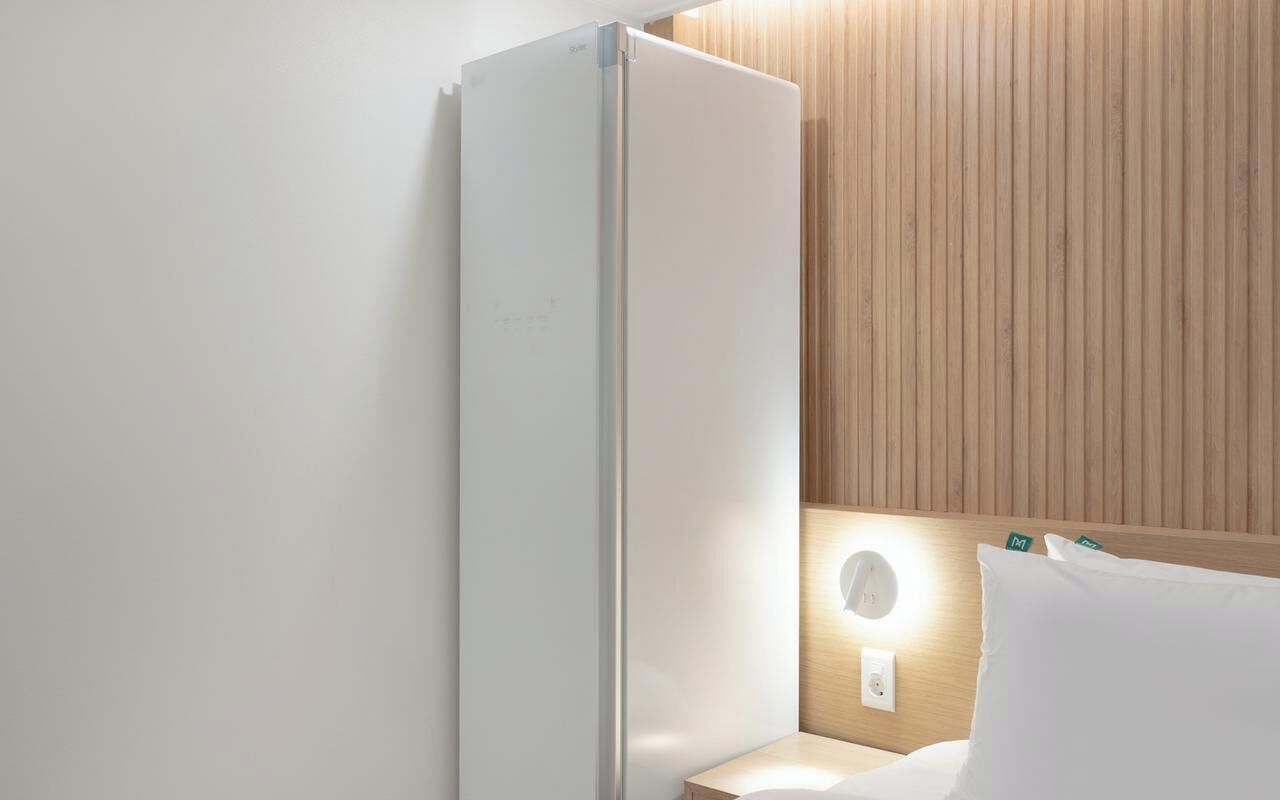 A hotel room with a white wall, a wooden panel wall, a bedside table with a lamp, and a white pillow.