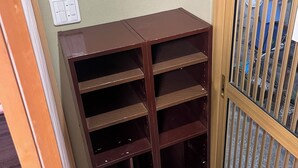 A corner room with a wooden bookshelf, a door with a metal grill, and a wall switch.