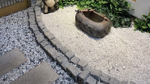 A stone pathway leading to a door with vertical blinds, a decorative stone basin, and a small statue.