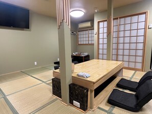 Una habitación con suelo de tatami, una mesa baja, sillas negras, un televisor montado en la pared y un panel corredero shoji.
