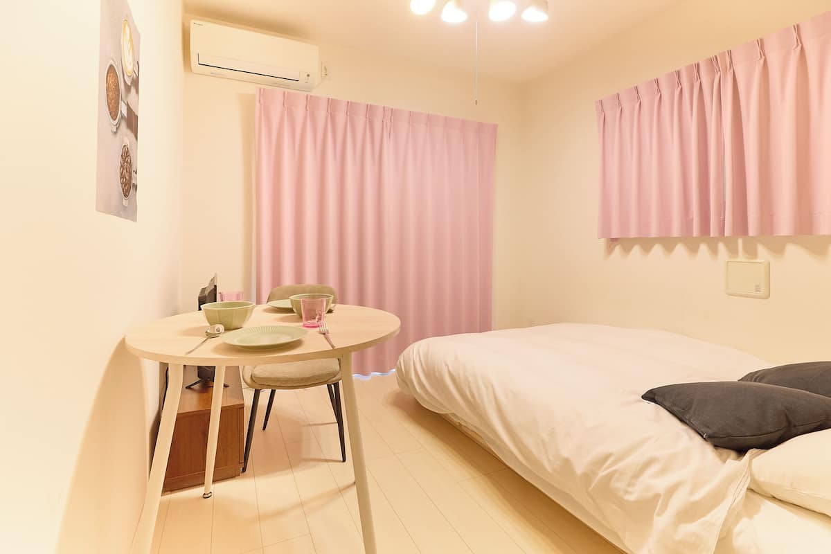 Quarto com cama, uma pequena mesa com cadeira, janela com cortinas rosa e ar condicionado.