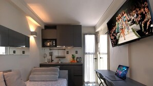 Quarto de hotel pequeno, com cama, escrivaninha, televisão e cozinha compacta.