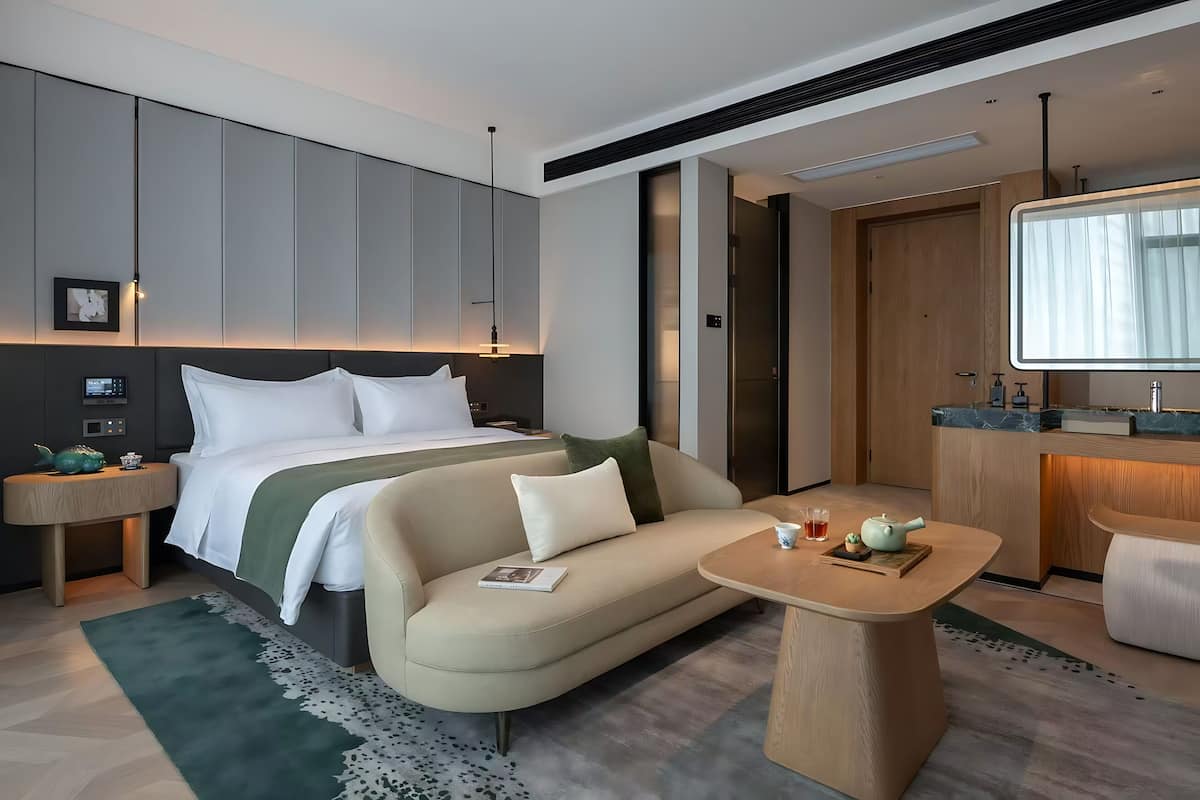 A modern hotel room with a bed, sofa, bedside table, and a small dining area.
