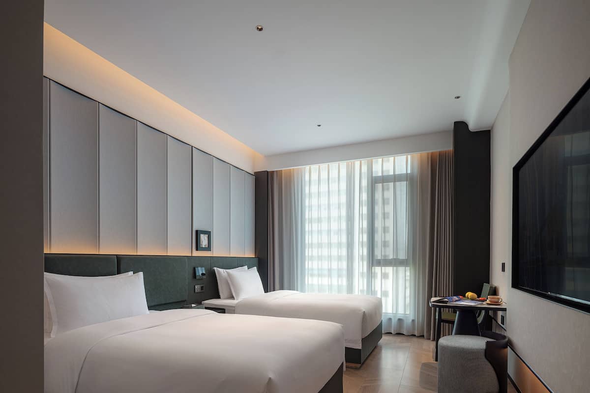 A hotel room with two beds, a desk, and a large window.