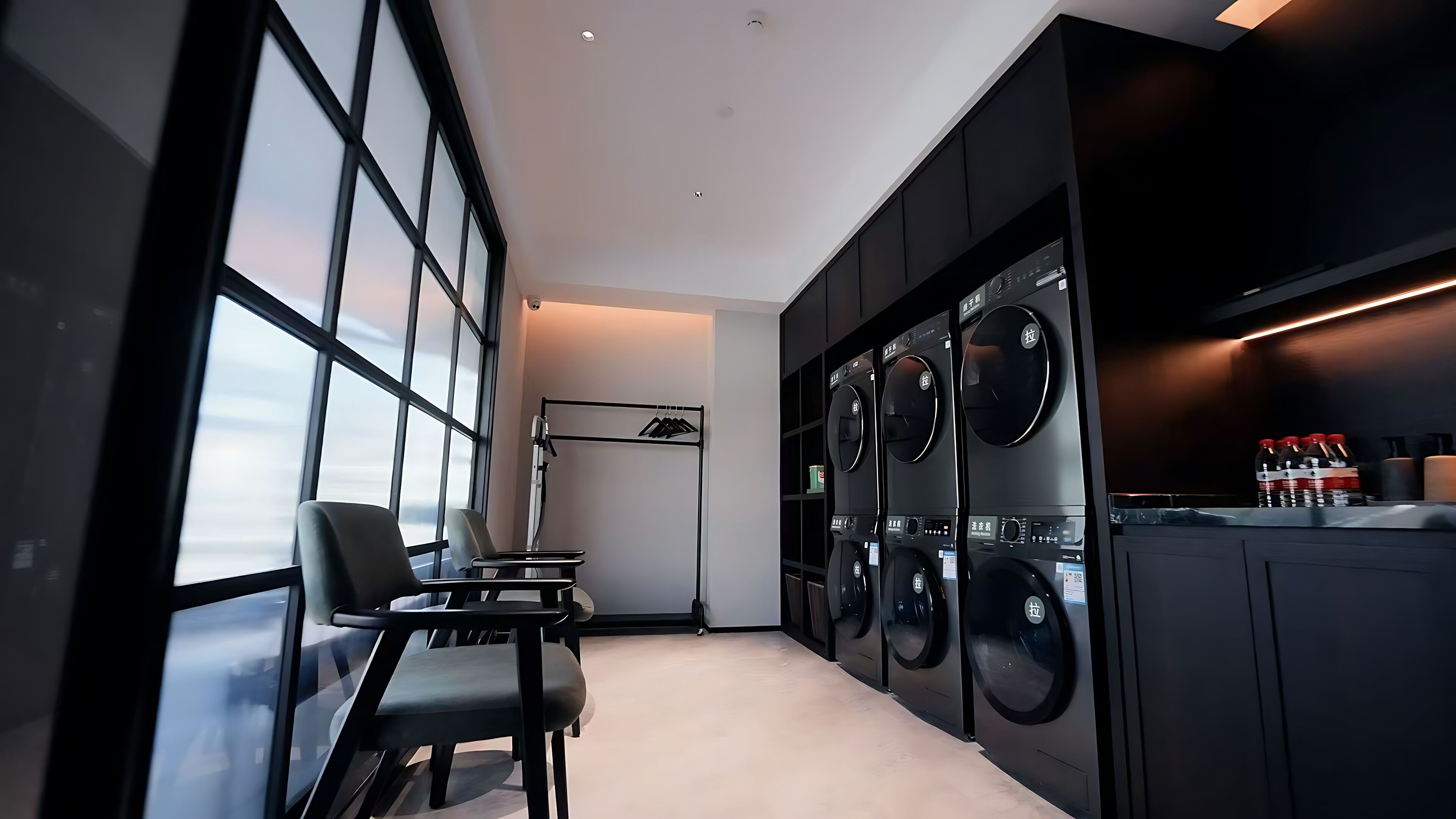 A modern laundry room with large windows, a seating area, and multiple washing machines.