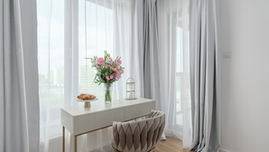 Une chambre moderne avec un bureau blanc, une chaise rembourrée, un vase de fleurs et des rideaux légers.