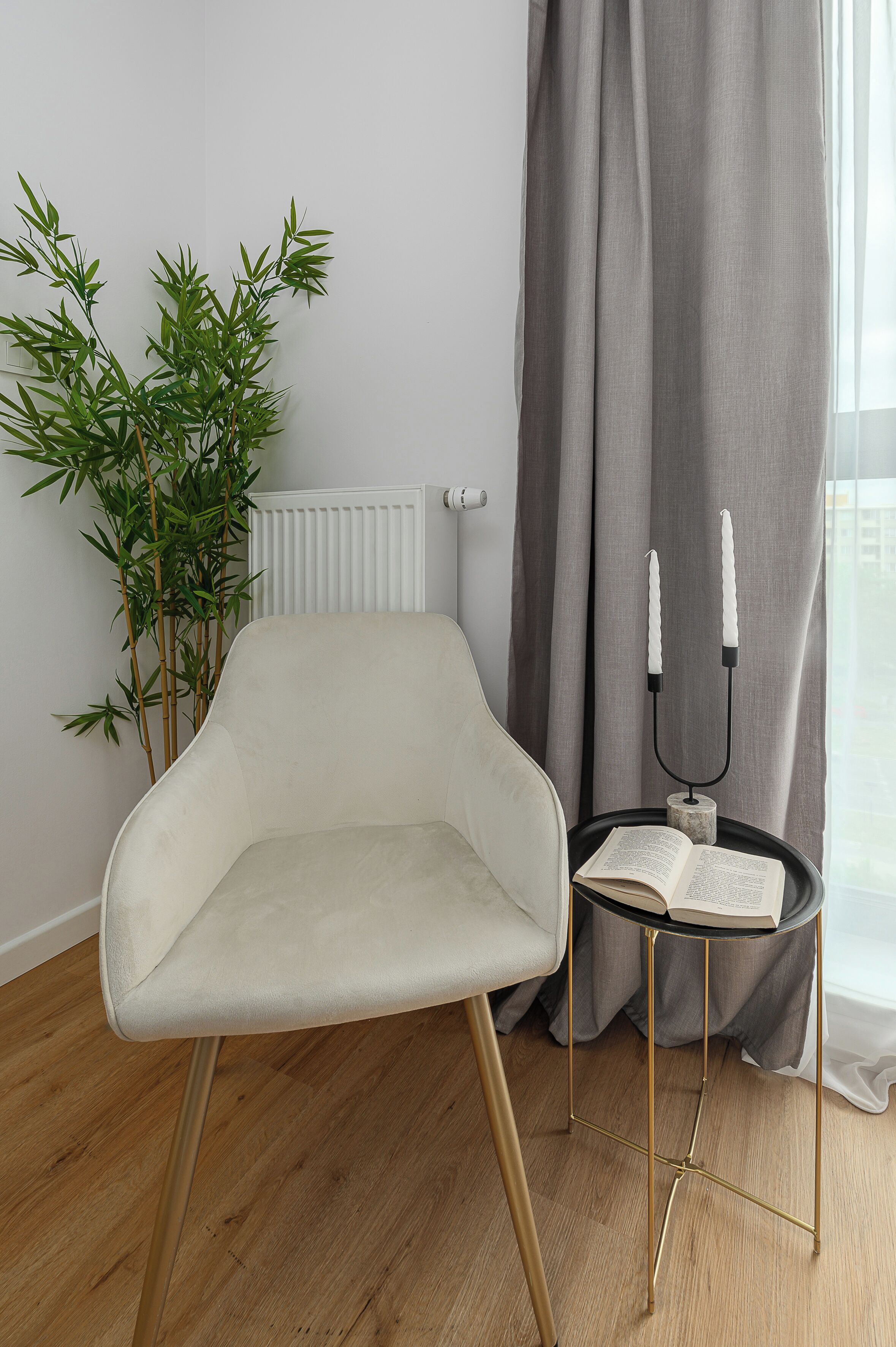 Een moderne kamer met een witte stoel, een klein rond tafeltje met een boek, een bamboeplant en een radiator.