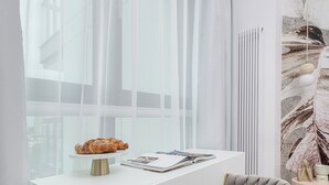 Une pièce moderne avec un bureau blanc, une chaise rembourrée de couleur grise, une fenêtre avec des rideaux légers et un élément décoratif mural.