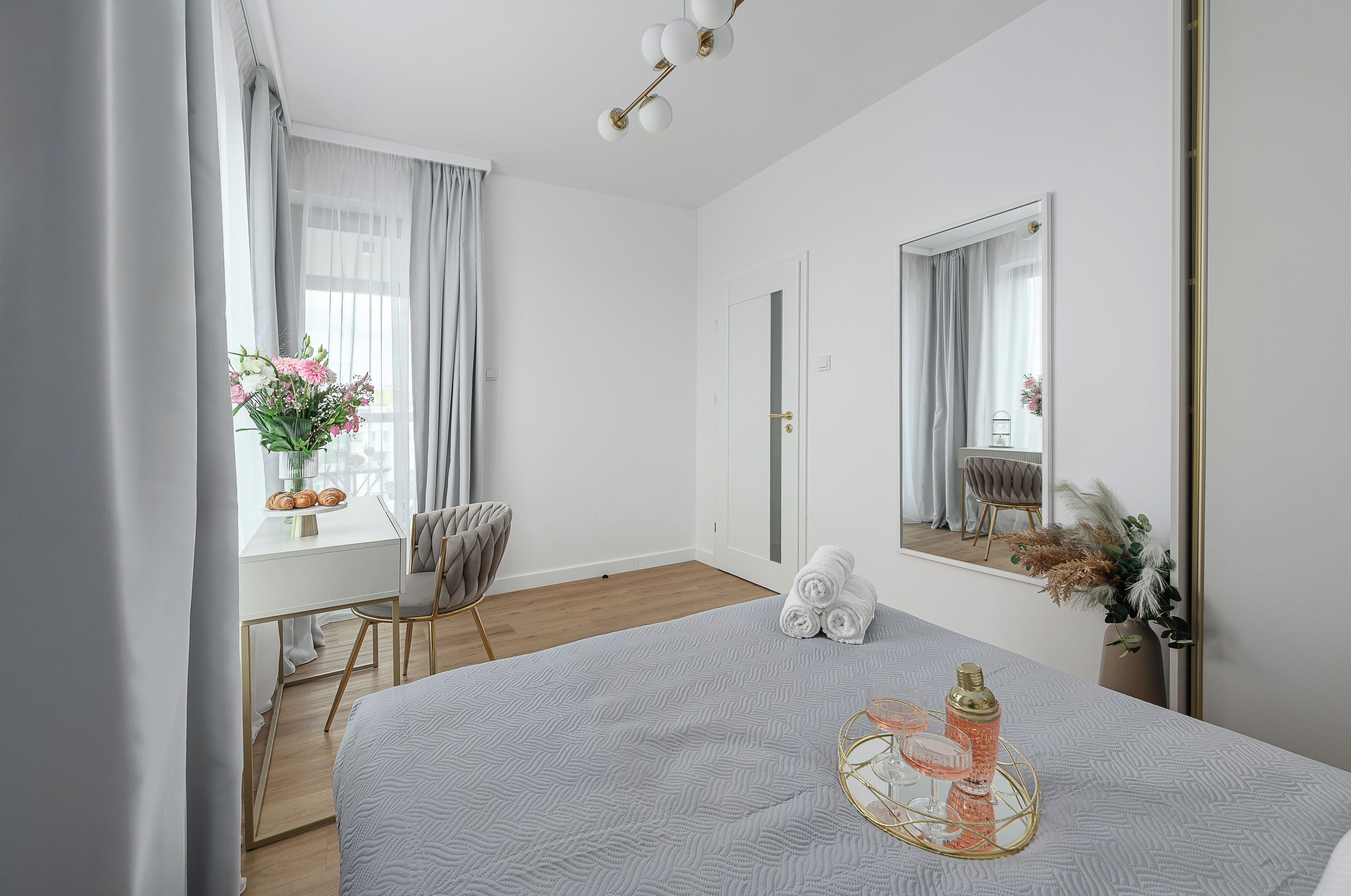 A bedroom with a bed, a desk, a chair, a window with curtains, a mirror, and a vase with flowers.