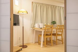 Ein kleines Zimmer mit Schreibtisch, Stuhl, Lampe und einem Fenster mit Vorhängen.