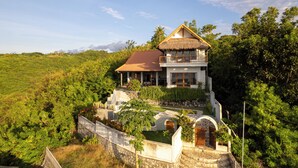 A two-story house with a thatched roof, surrounded by lush greenery and a stone wall.
