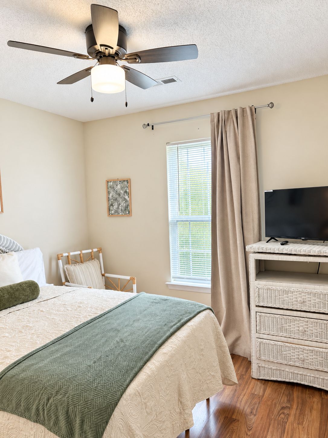 A bedroom with a bed, a ceiling fan, a television on a wicker stand, a window with curtains, and a framed picture on the wall.