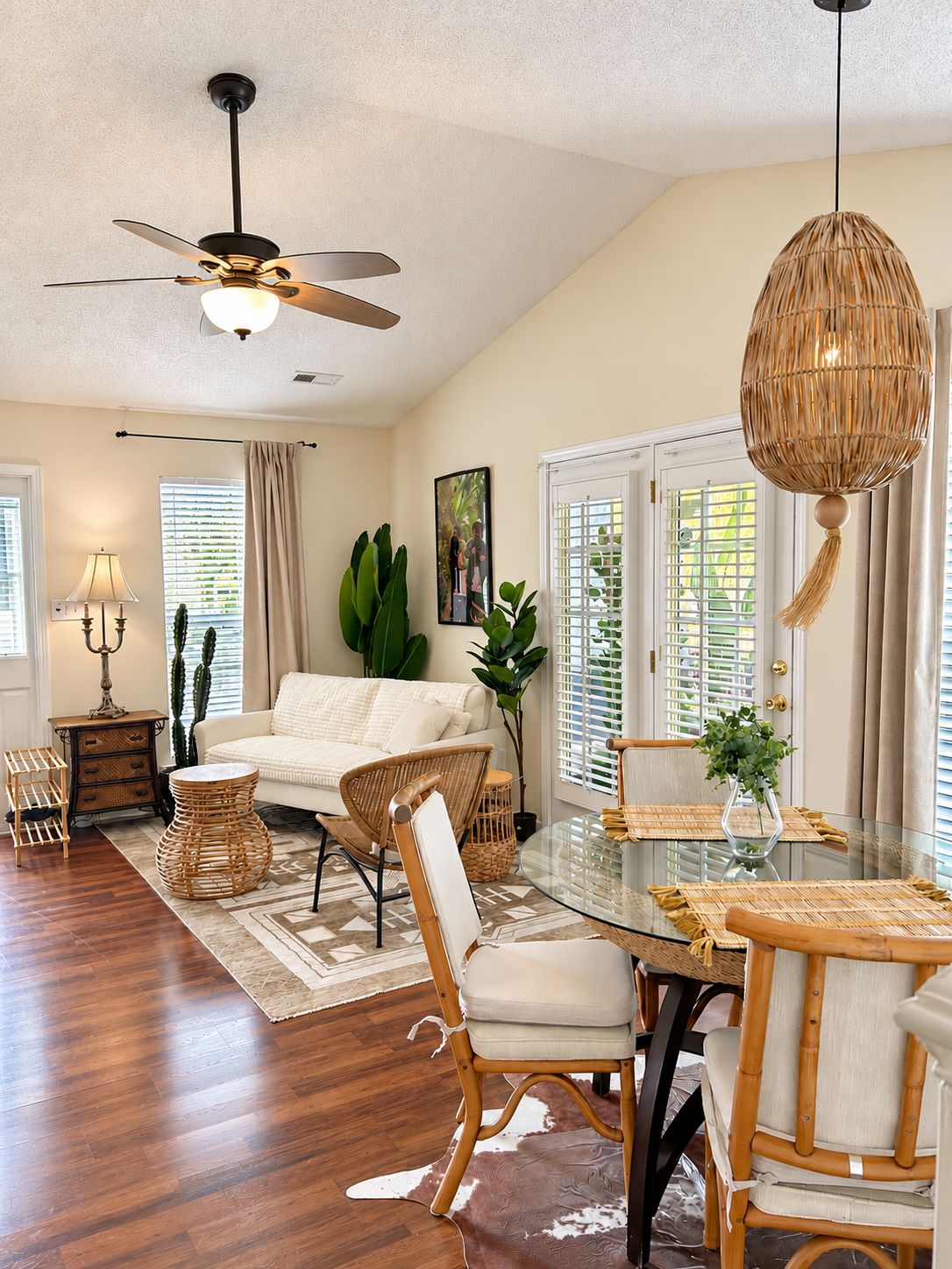 A spacious living room with a ceiling fan, a glass dining table with a woven chair, and a large window with blinds.
