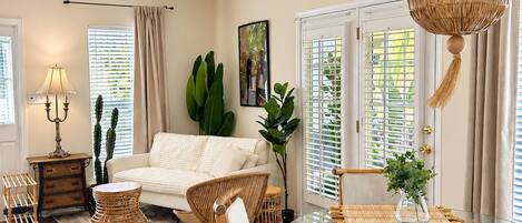 A spacious living room with a ceiling fan, a glass dining table with a woven chair, and a large window with blinds.