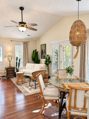 A spacious living room with a ceiling fan, a glass dining table with a woven chair, and a large window with blinds. - Tropical bungalow with screened porch (Charlotte)