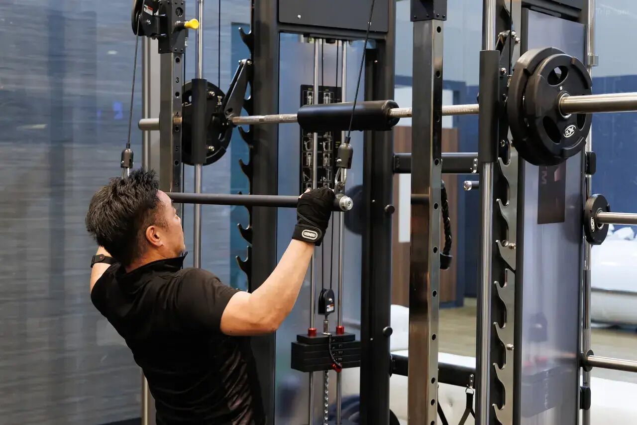 A person using a weightlifting machine in a gym with a large window in the background.