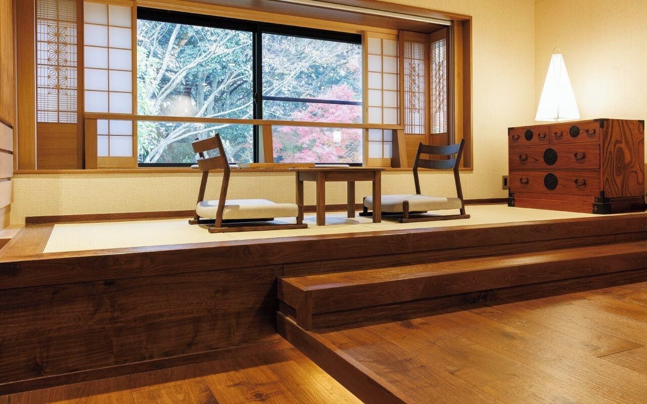 A traditional Japanese room with tatami flooring, a low table with chairs, a wooden cabinet, and a window with a view of trees.