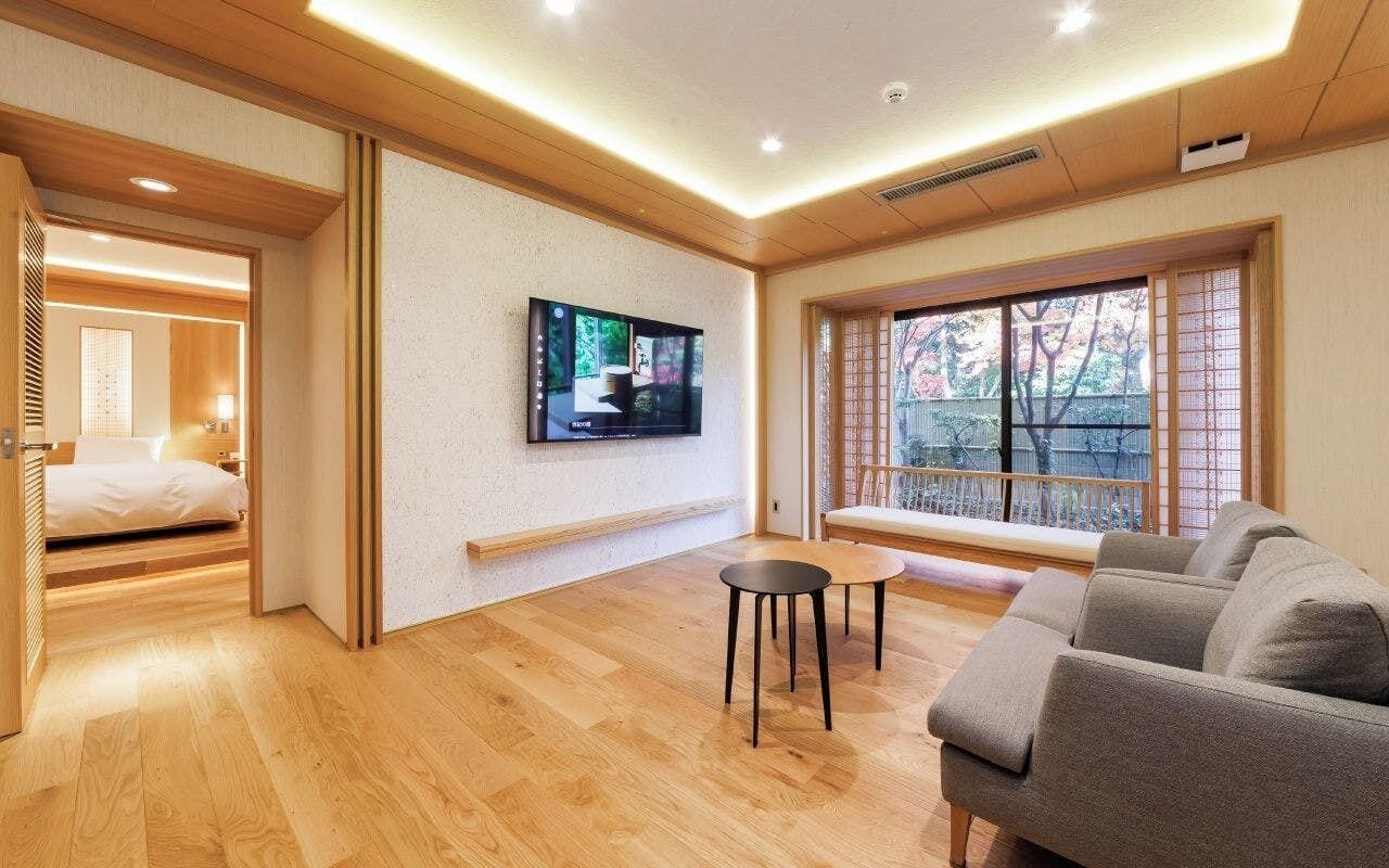 A modern hotel room with a wooden floor, a sofa, a TV mounted on the wall, and a window with a view of trees.