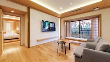 A modern hotel room with a wooden floor, a sofa, a TV mounted on the wall, and a window with a view of trees.