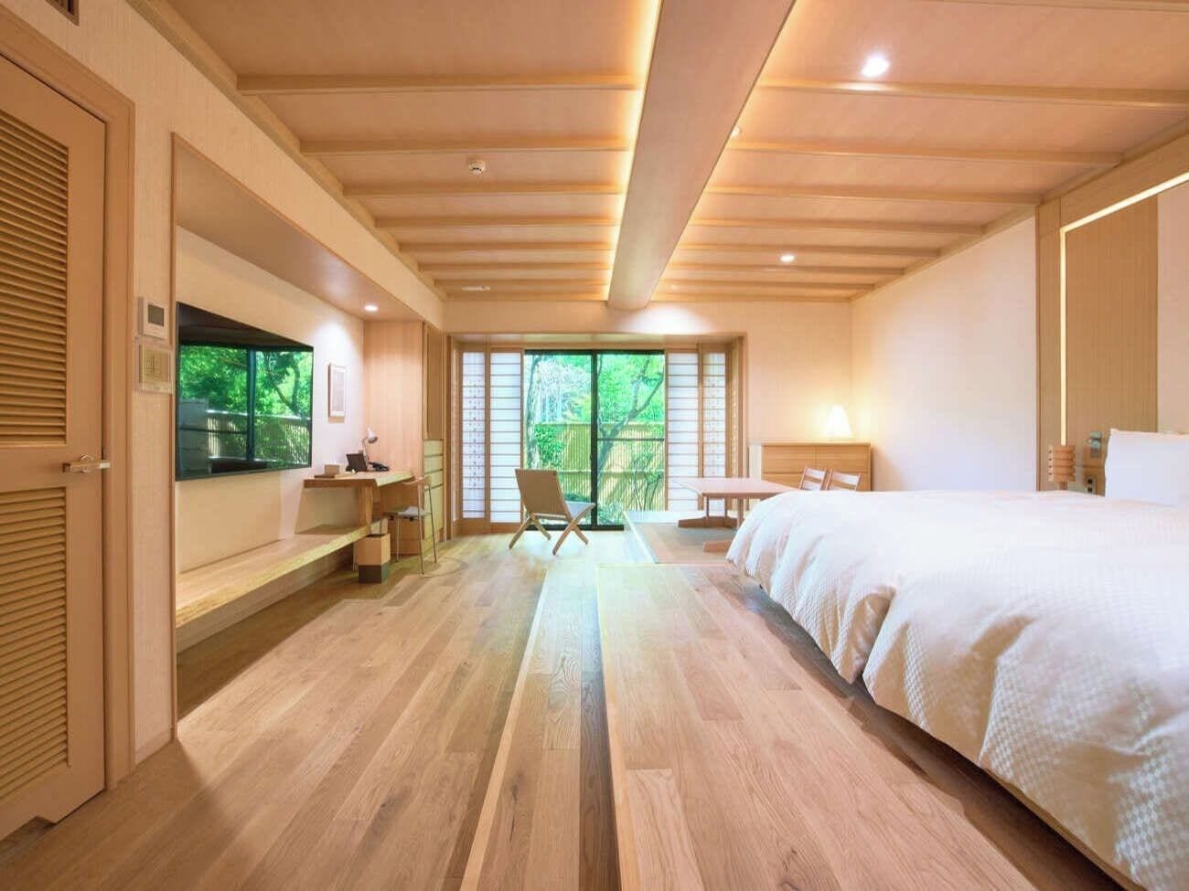 A hotel room with a wooden floor, a bed, a desk, and a chair. There is a window with a view of greenery outside.