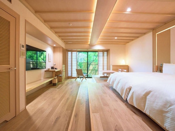 Habitación de hotel con piso de madera, una cama, un escritorio y una silla. Se ve un ventanal con vista a zonas verdes.
