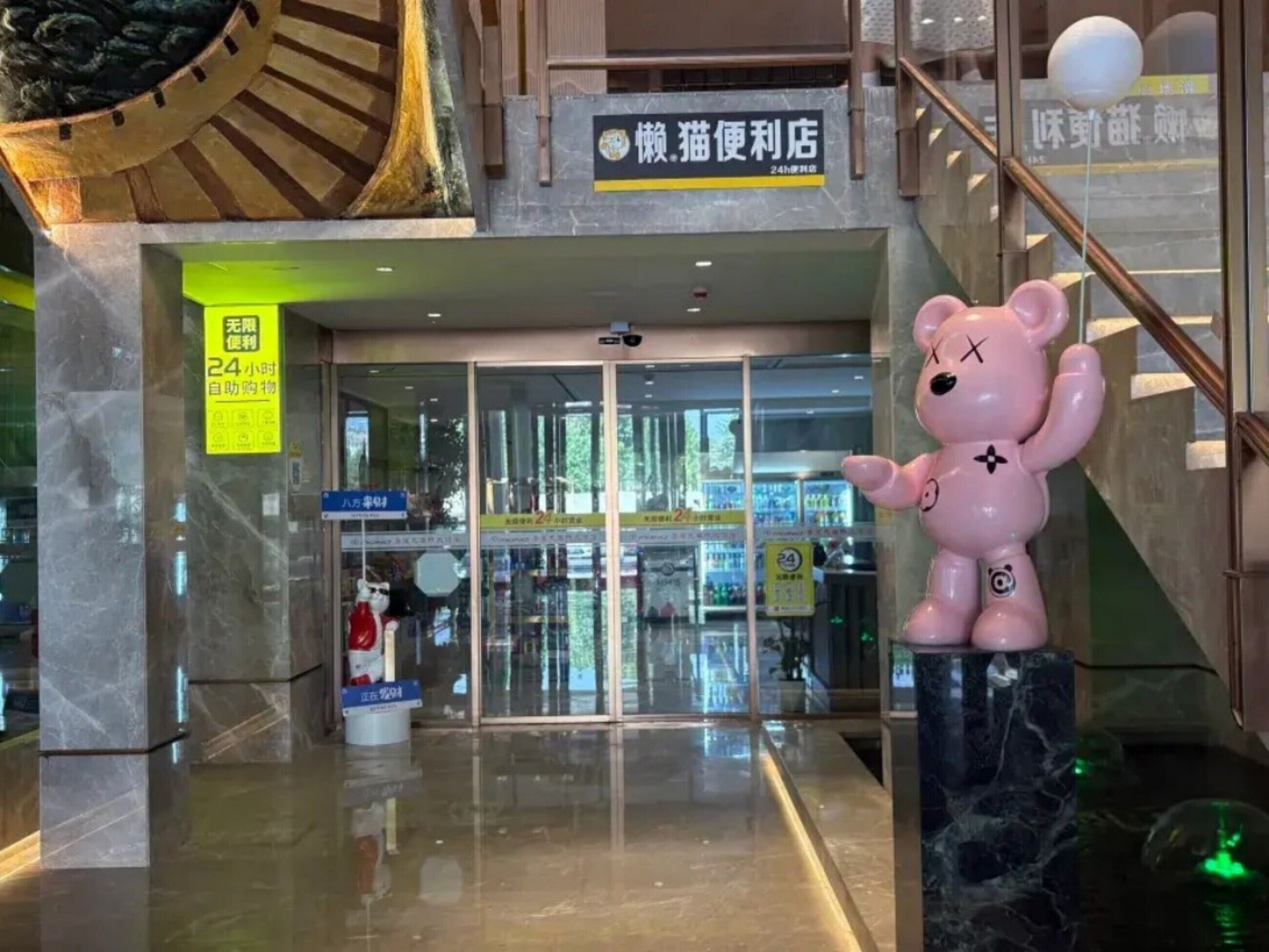Entrada a un centro comercial con una tienda de conveniencia abierta las 24 horas, una gran estatua de un oso rosa y un extintor.