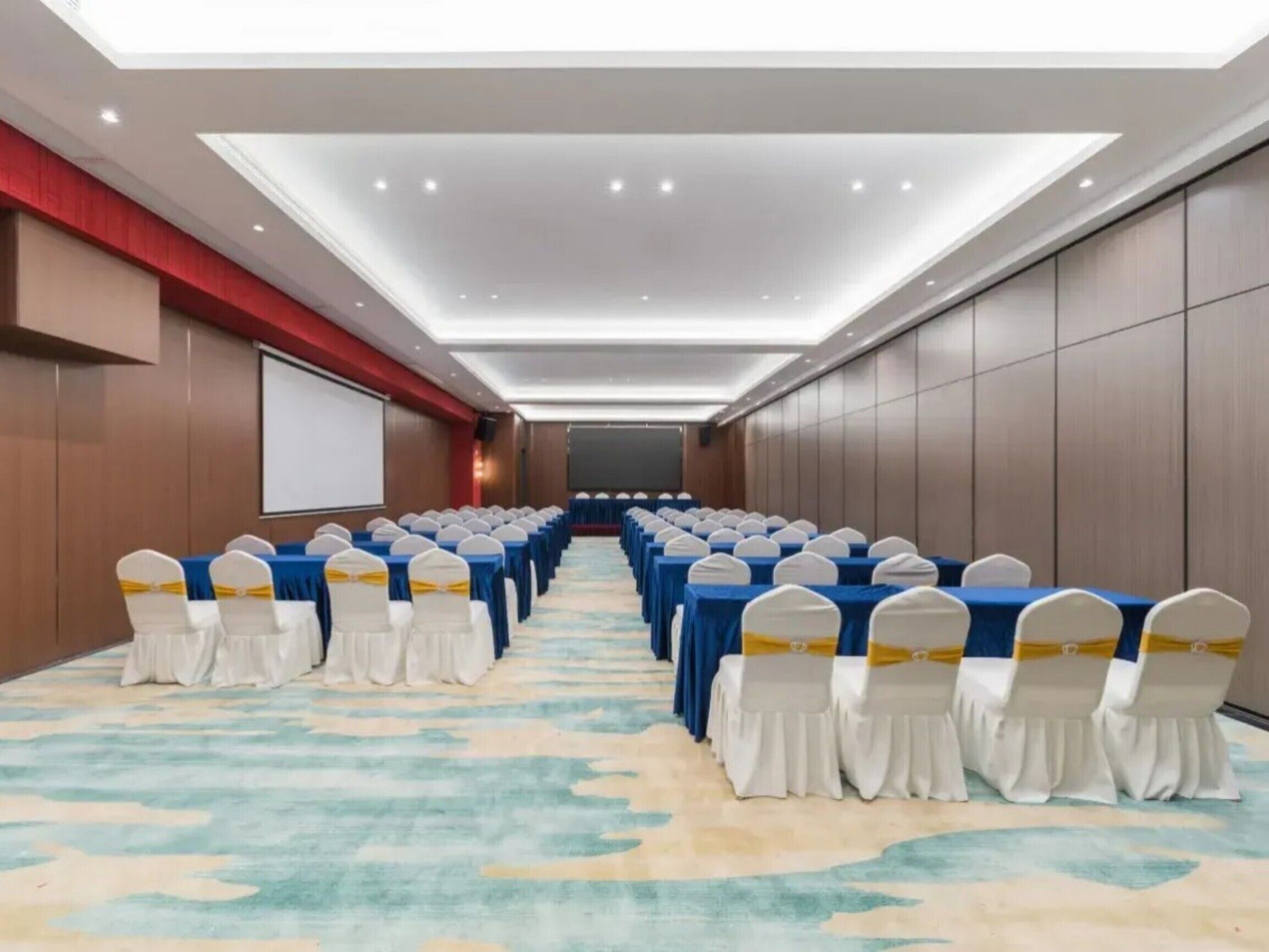 A conference room with rows of tables and chairs, a projector screen, and a carpeted floor.