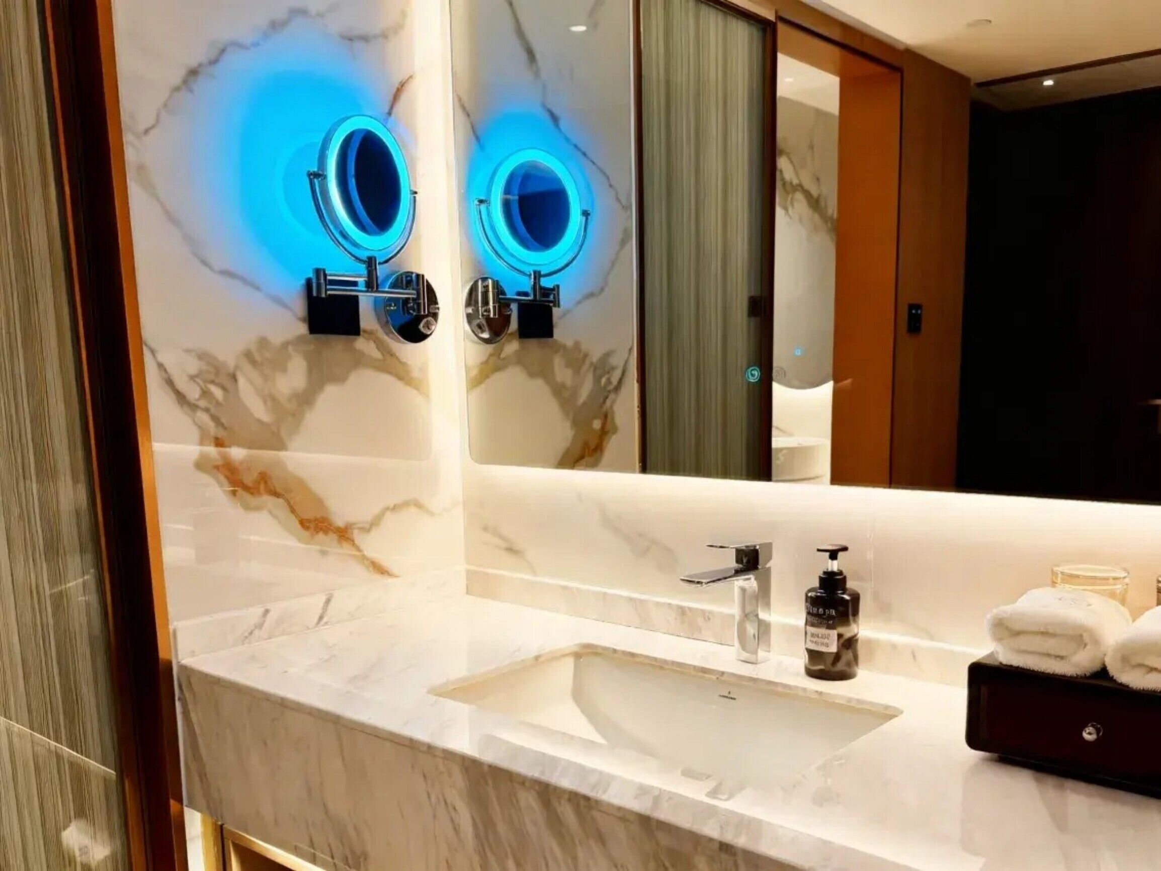 A bathroom with a marble vanity, a sink, two illuminated mirrors, a bottle of shampoo, and towels.